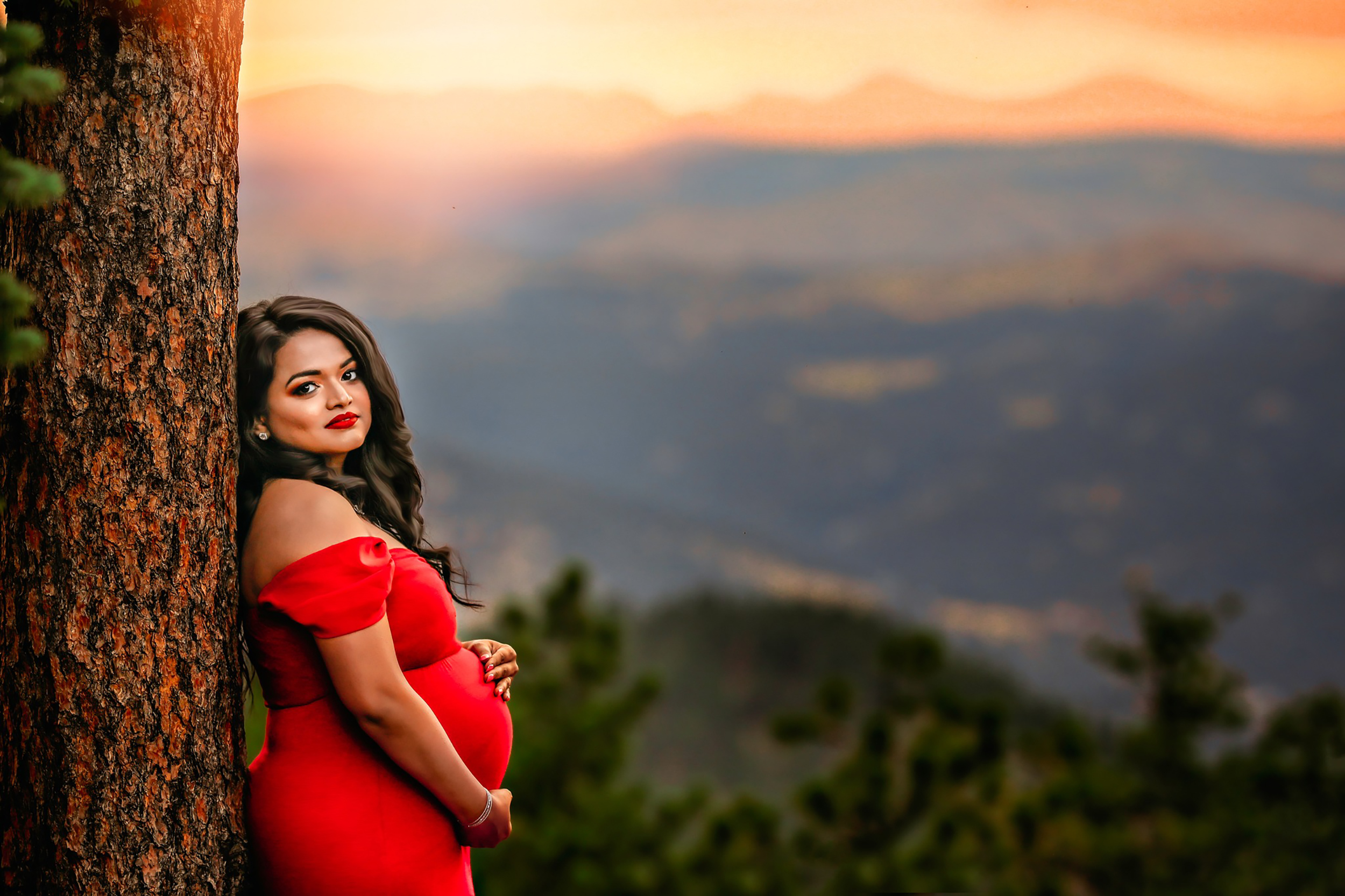 A mother to be in a red maternity gown leans against a tree on a mountaintop with mountains in the backgrround