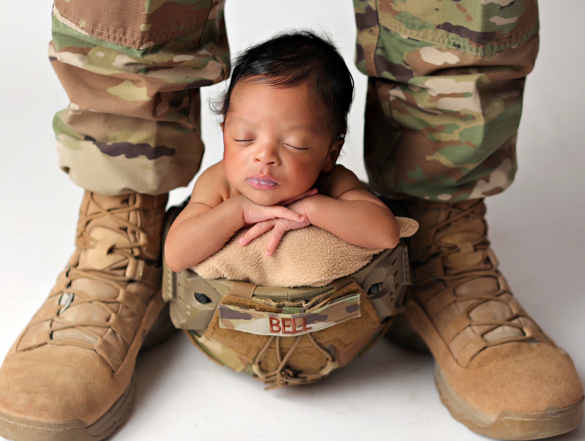 Newborn posing between dad's legs in dad's military helmet.