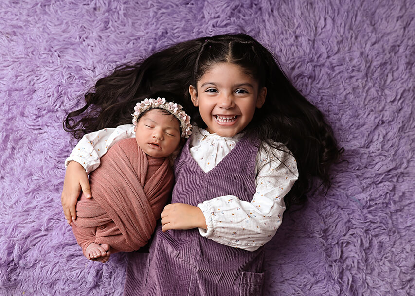 Newborn baby and sister posing together on purple blanket at their Denver newborn photos session.