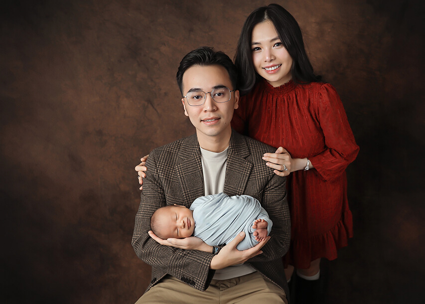 Mom and Dad posing in front of brown backdrop holding their sleeping newborn baby. 