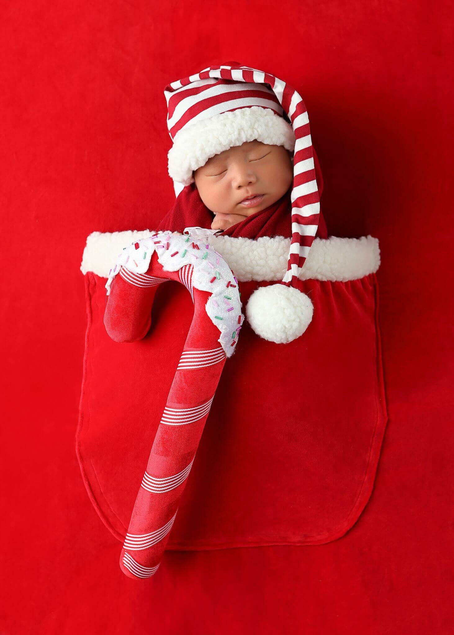 A cute newborn sleeping in a Santa Claus sack and holding a candy cane in his is one of our best holiday-themed photoshoot ideas.