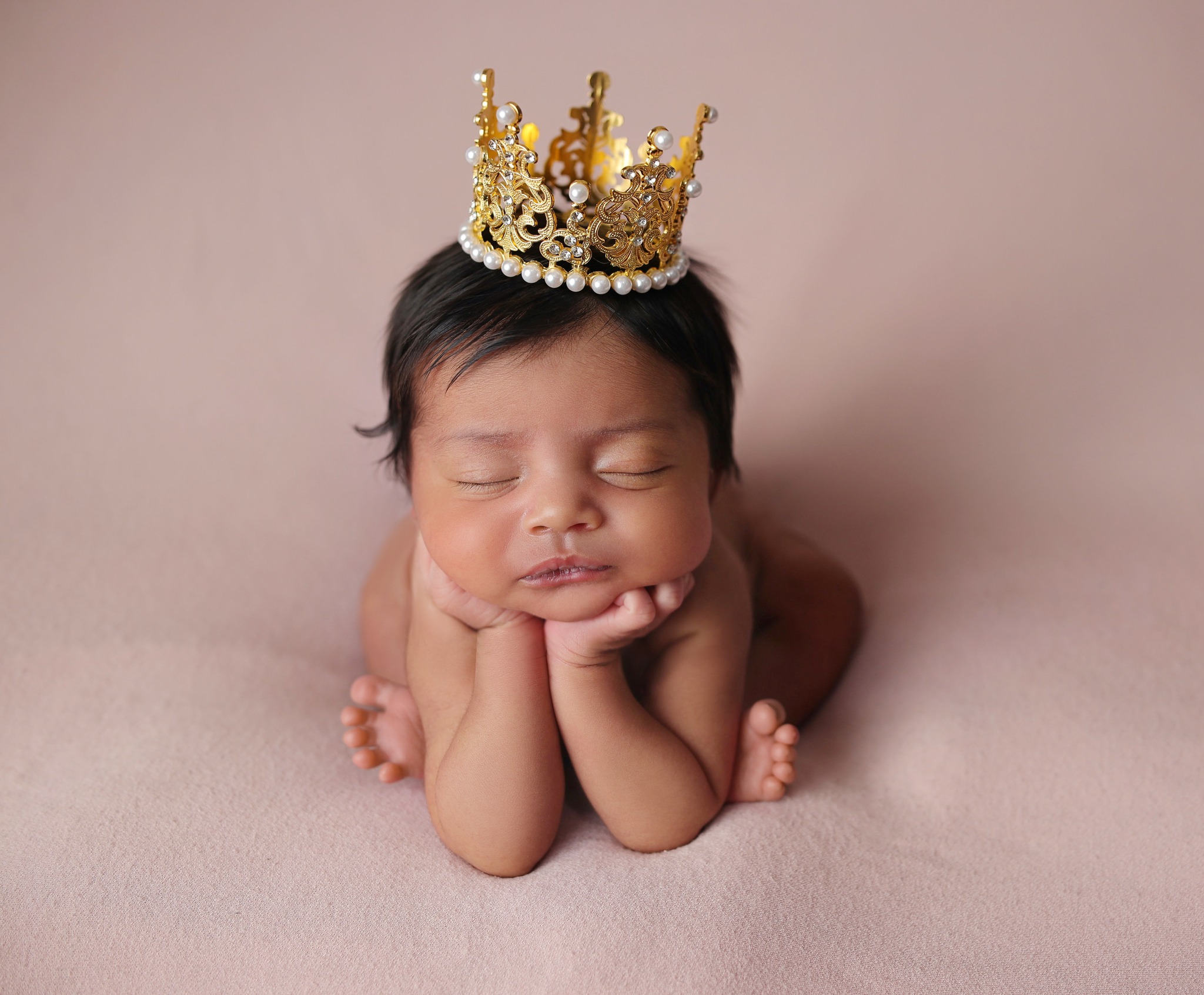 Sleeping newborn in frog pose wearing a gold crown on pink beanbag blanket. 