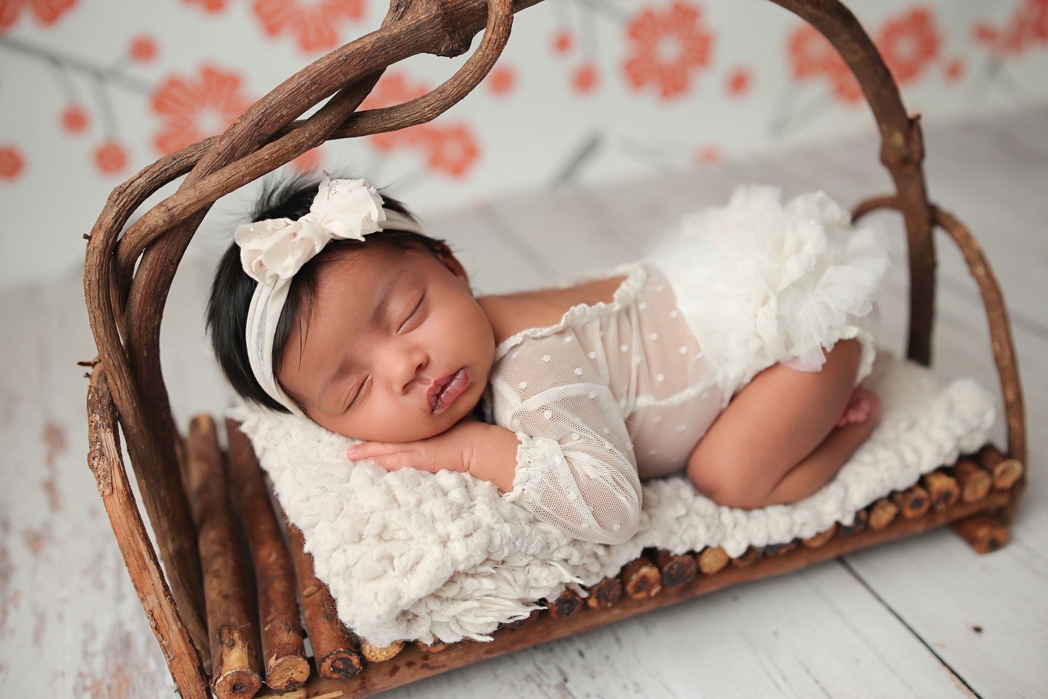 Newborn baby sleeping on wicker bed in white lacey outfit.